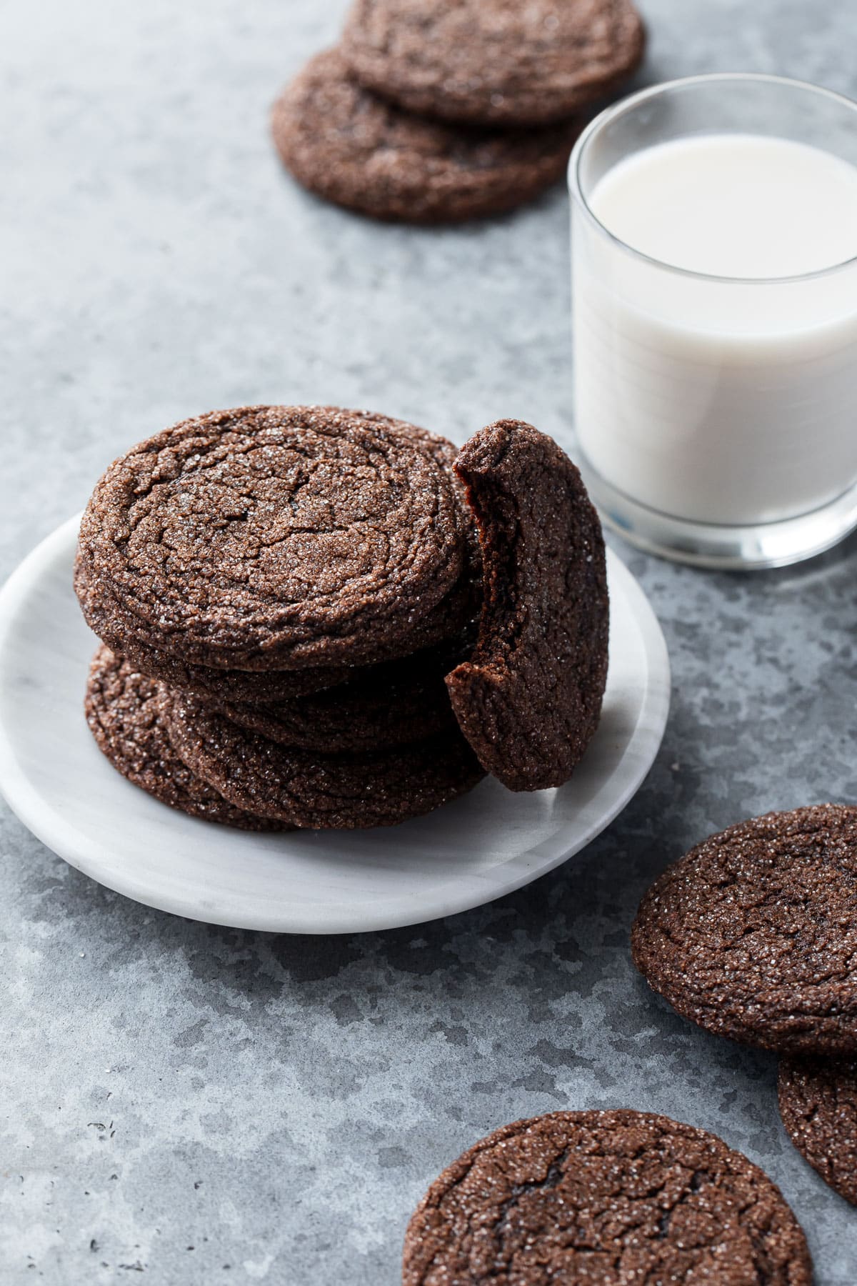 Stack of Chewy Chocolate Molasses Cookies on a plate with one bitten cookie resting on its side, glass of milk and a few more cookies scattered around.