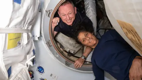 NASA Butch Wilmore looking through a hatch on the international space station and Suni Williams lying just outside. Both are smiling and Suni's hair is waving in the zero gravity environment