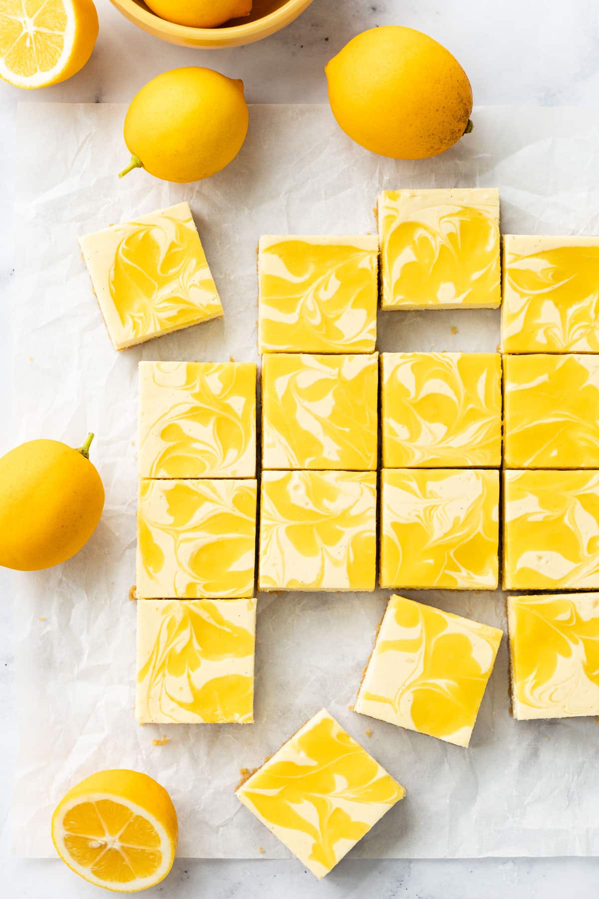 Overhead, squares of Meyer Lemon Cheesecake Bars with swirls of bright yellow lemon curd, on a piece of crinkled parchment with a few whole and cut lemons.