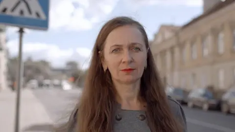 BBC A woman with long brown hair stands in a street and stares at a camera