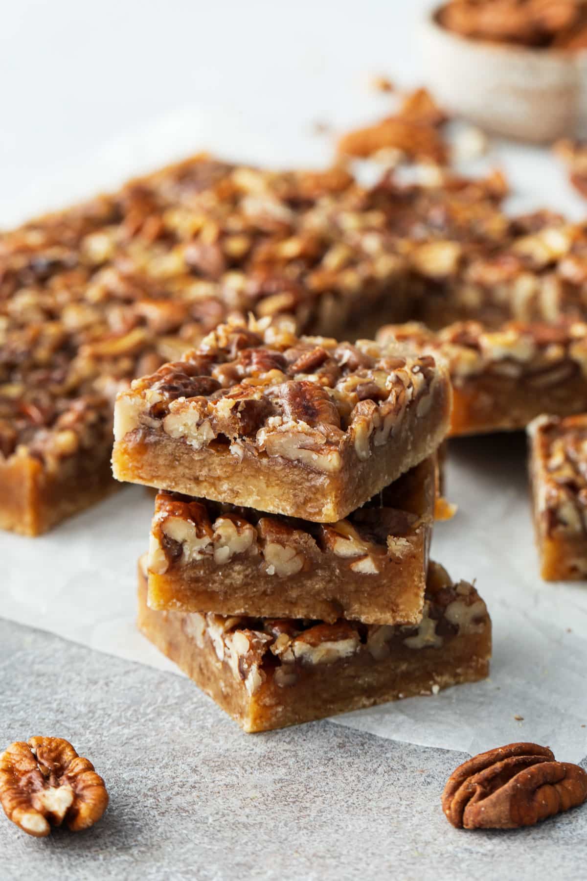 Stack of three square cut Pecan Pie Shortbread Cookie Bars on parchment, with the rest of the pan of bars in the background.