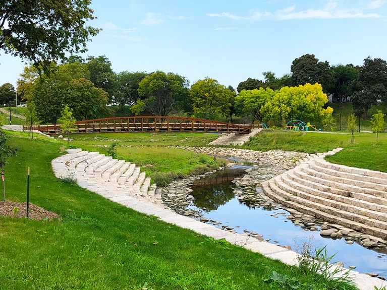 At the top is blue sky, below is a row of green trees, and below that a bridge crossing a river which can be seen in the foreground. The river is edged by stones, and has some steps to the left and right. Grassy banks surround the river.