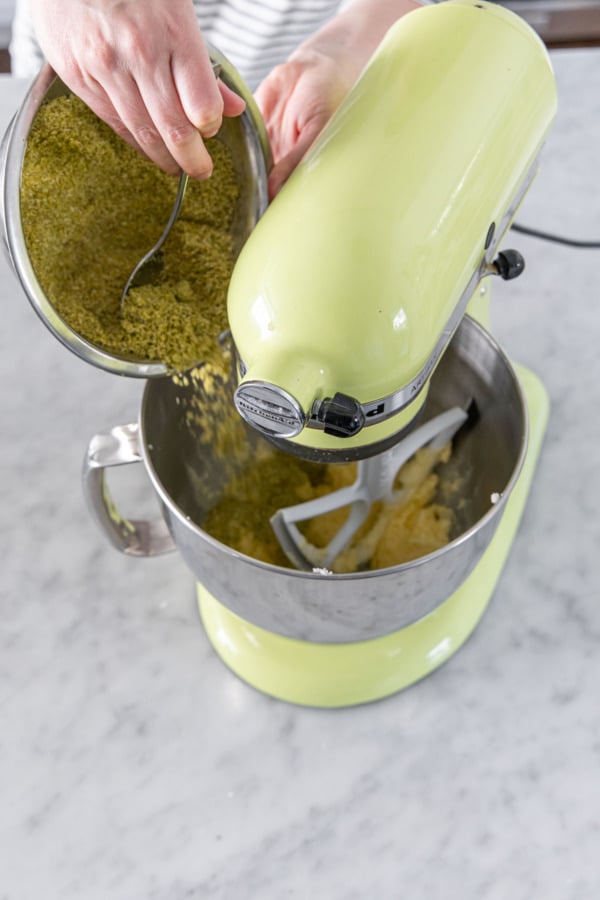 Adding half of pistachio flour mixture to mixing bowl with frangipane filling.