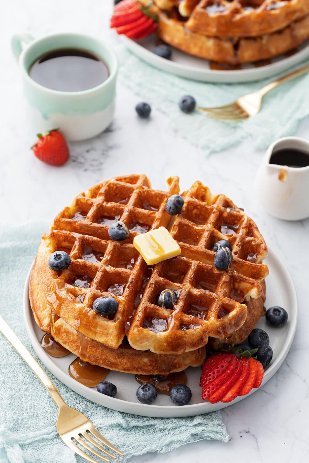 Plates with stacks of golden brown sourdough Belgian waffles topped with berries, a pat of butter and drizzled maple syrup, cup of coffee in the background.